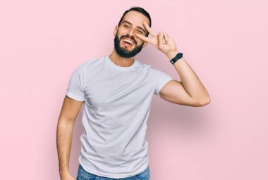 Sakallı, günlük beyaz tişörtlü genç adam barış sembolü yapıyor. Yüzünde parmaklar, gülümseyerek zaferi gösteriyor. 