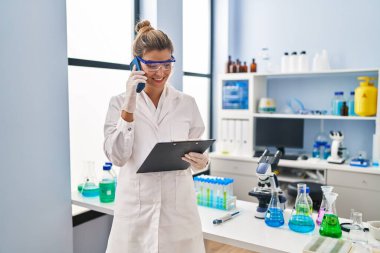 Bilim adamı üniforması giyen genç sarışın kadın laboratuvarda akıllı telefondan konuşuyor.