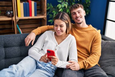 Genç adam ve kadın çift akıllı telefon kullanarak evdeki kanepede oturuyorlar.