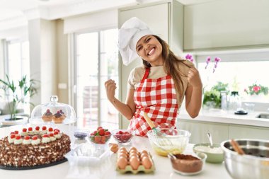 Güzel, genç, esmer pasta şefi kadın mutfakta hamur işleri pişiriyor. Çok mutlu ve heyecanlı, kolları havada, gülümseyerek ve başarı için çığlık atarak kazanan jestler yapıyor. kutlama konsepti. 
