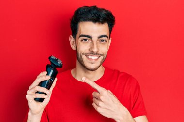 Elektrikli tıraş makinesi tutan genç İspanyol adam gülümsüyor mutlu bir şekilde eli ve parmağı işaret ediyor. 