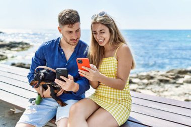 Akıllı telefon kullanan beyaz bir çift sahilde köpeğiyle bankta oturuyor..