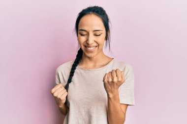 Gündelik beyaz tişört giyen genç İspanyol kız şaşkınlık ve şaşkınlık içinde kollarını kaldırarak ve gözlerini kapayarak başarıyı kutluyor. 