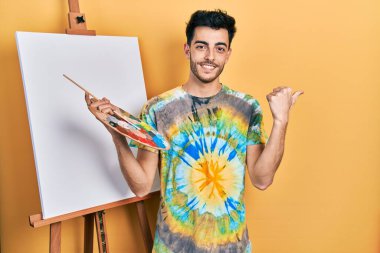 İspanyol asıllı genç adam paletle resim çiziyor, resim sehpası, neşeli duruyor, yüzünde bir gülümsemeyle, mutlu ve doğal bir ifadeyle elini ve parmağını yana kaldırıyor. 