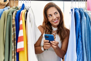 Genç Latin bayan müşteri akıllı telefon kullanıyor giyim mağazasında elbise askılığında beliriyor