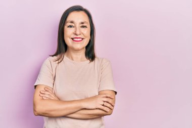 Orta yaşlı, kollarını kavuşturup gülümseyen, yüzünde mutlu ve havalı bir gülümsemeyle İspanyol bir kadın. diş gösterme. 