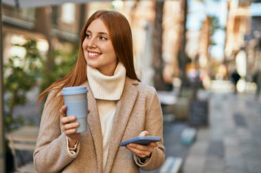 Genç kızıl saçlı kız akıllı telefon kullanıyor. Şehirde kahve içiyor..