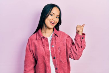 Güzel İspanyol bir kadın, burun piercingli, sıradan pembe bir ceket giyiyor, gülümsüyor, gülümsüyor ve baş parmağıyla yan tarafa bakıyor.. 