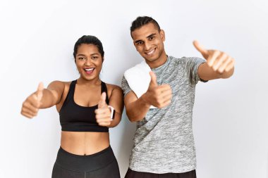 Spor kıyafetleri giyen genç bir çift izole edilmiş arka planda durup el hareketleriyle olumlu jest yapmayı onaylıyor, gülümsüyor ve başarı için seviniyor. kazanan jest. 