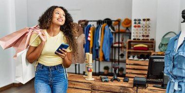 Genç Latin müşteri kadın gülümseyerek alışveriş torbaları tutuyor ve giyim mağazasında akıllı telefon kullanıyor..