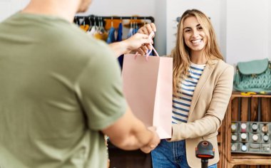 Genç İspanyol esnaf gülümseyerek giyim mağazasındaki müşteriye alışveriş çantası veriyor..