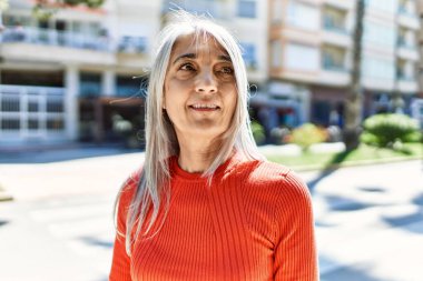 Orta yaşlı, kır saçlı, gülümseyen bir kadın şehirde mutlu mesut duruyor..