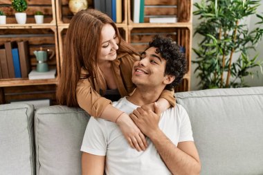 Mutlu gülümseyen ve sarılan genç çift evdeki kanepede oturuyor..