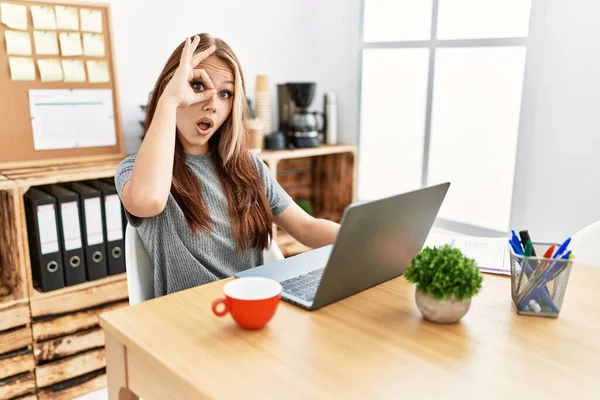 Young brunette woman working at the office with laptop doing ok gesture shocked with surprised face, eye looking through fingers. unbelieving expression.