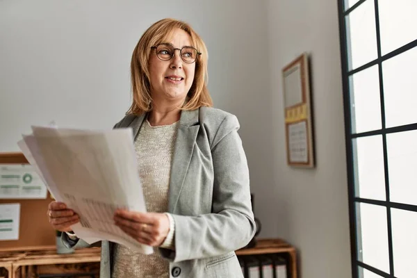 Middle age businesswoman smiling happy holding paperwork standing at the office.