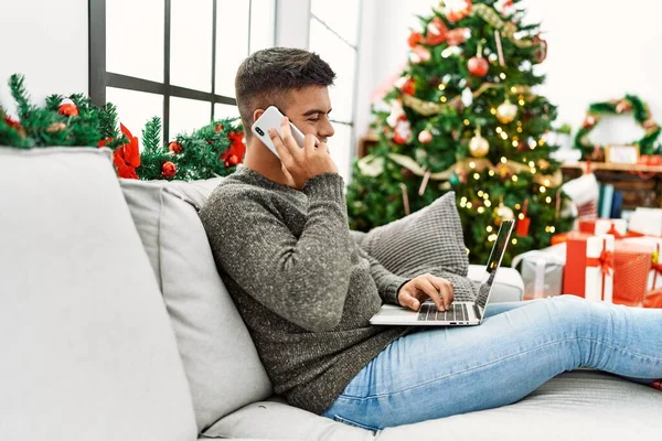 年轻的他的惊慌失措的男人在家里坐在圣诞树旁的沙发上 用笔记本电脑和智能手机聊天 — 图库照片