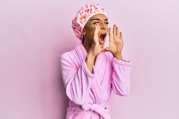 Hispanic man wearing make up wearing shower towel cap and bathrobe shouting angry out loud with hands over mouth
