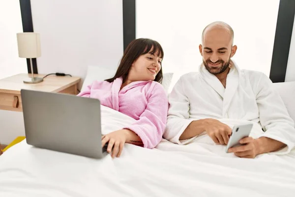 他的年轻夫妇坐在卧室的床上 他们使用笔记本电脑和智能手机 — 图库照片