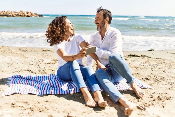 中年时 他的一对惊慌失措的夫妇坐在沙滩上的毛巾上 用智能手机 — 图库照片