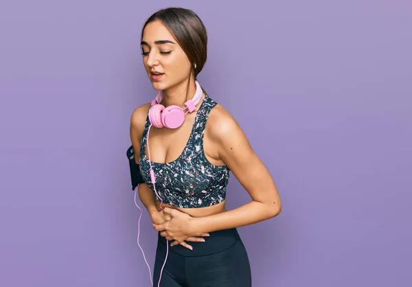 Beautiful brunette woman wearing gym clothes and using headphones with hand on stomach because nausea, painful disease feeling unwell. ache concept.