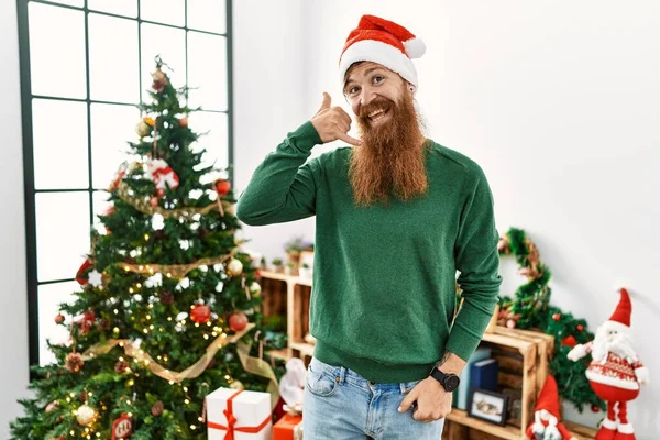 红头发的男人留着长胡子 头戴圣诞帽 站在圣诞树旁笑着用手和手指做着电话手势 就像在讲电话一样 交流概念 — 图库照片