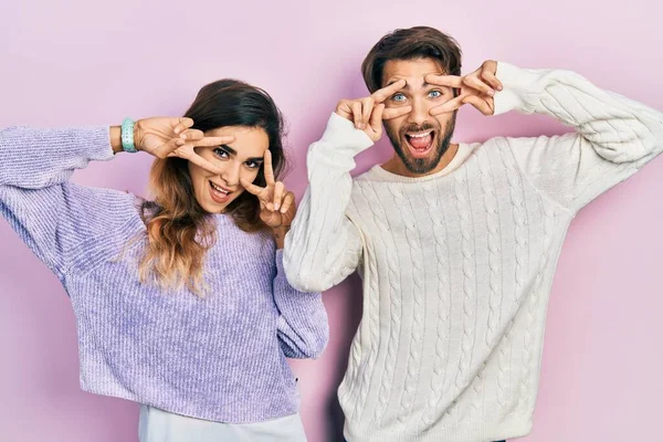 若いヒスパニック系のカップルは 顔の上に指で平和のシンボルを行うカジュアルな服を着て 明るい勝利を示す笑顔 — ストック写真