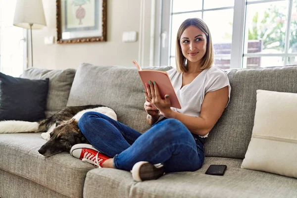 年轻的高加索女孩高兴地坐在沙发上 带着狗在家里看书 — 图库照片