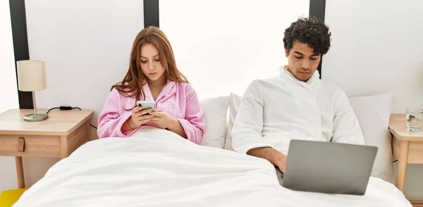 有严肃表情的年轻夫妇在卧室里使用笔记本电脑和智能手机 — 图库照片