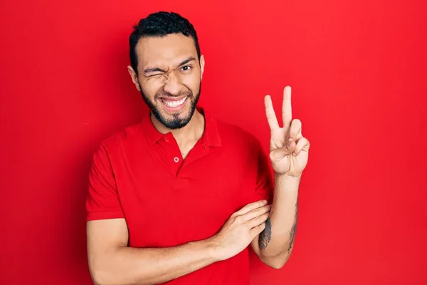 カジュアルな赤いTシャツを着て髭を生やしたヒスパニック系の男が カメラで勝利のサインをして 幸せな顔をして笑っている — ストック写真