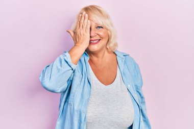 Orta yaşlı sarışın kadın gündelik kıyafetler giyiyor. Bir gözünü eliyle kapatıyor. Yüzünde kendine güvenen bir gülümseme ve sürpriz bir duygu var.. 