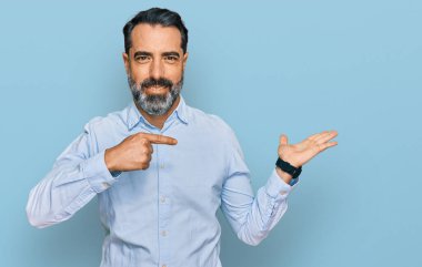 Orta yaşlı, sakallı, iş gömleği giyen bir adam kameraya hayret ediyor ve gülümsüyor.. 