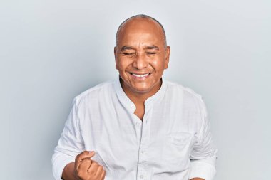 Orta yaşlı, gündelik kıyafetler giyen, mutlu ve heyecanlı bir Latin adam kolları havada, gülümseyen ve başarı için bağıran, kazanan bir jest yapıyor. kutlama konsepti. 