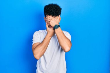 Sakallı, beyaz tişörtlü, gözleri ve ağzı elleri kapalı, şaşırmış ve şok olmuş genç bir Arap. duygu gizleme 