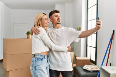 Genç beyaz çift mutlu bir şekilde gülümsüyor. Yeni evlerinde akıllı telefonların yanında selfie çekiyorlar..