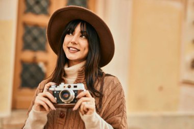Kışlık şapkalı esmer kadın şehrin dışında klasik kamera kullanarak gülümsüyor.