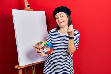 Orta yaşlı İspanyol kadın paletle resim çiziyor, sehpa boyuyor, elleriyle para hareketi yapıyor, maaş istiyor, milyonerlik işi istiyor. 