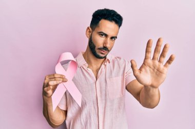 Genç İspanyol adam elinde pembe kanser kurdelesi ile elinde dur işareti ile ciddi ve kendinden emin bir ifadeyle savunma hareketi yapıyor. 