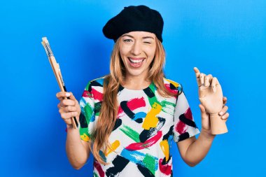 Küçük ahşap manikin ve boya fırçaları tutan genç beyaz kadın sanatçı kameraya seksi bir ifadeyle bakıyor, neşeli ve mutlu bir yüz.. 