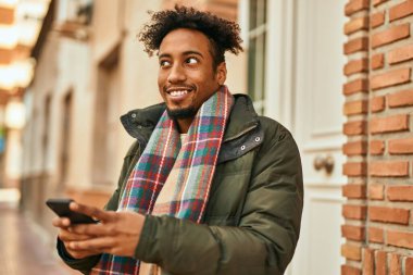 Genç Afrikalı Amerikalı adam şehirde akıllı telefon kullanarak mutlu bir şekilde gülümsüyor.
