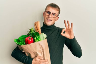 Kafkasyalı genç bir adam elinde poşetle market alışverişi yapıyor. Parmaklarıyla imza atıyor, gülümseyerek mükemmel bir sembol oluşturuyor. 