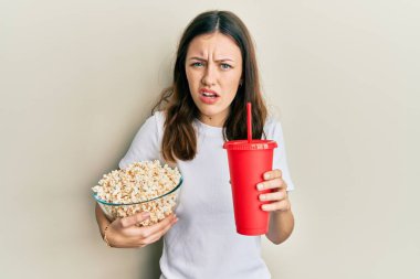 Genç esmer kadın patlamış mısır yiyor ve soda içiyor habersiz ve kafası karışık bir ifade. şüphe kavramı. 