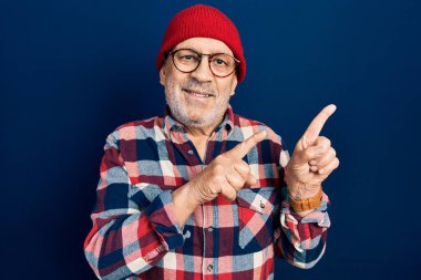 Yakışıklı, olgun, hippi görünümlü, yün şapkalı, gülümseyen ve kameraya bakan iki eli ve parmakları yandan gösteren bir adam.. 