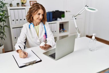 Orta yaşlı İspanyol kadın klinikte elinde haplarla doktor üniforması giyiyor.