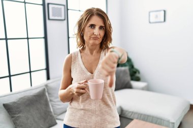 Orta yaşlı bir kadın evde kahve içerken mutsuz ve kızgın görünüyor. Reddedilmiş ve olumsuz bir hareket yapıyor. kötü ifade. 