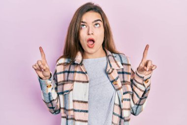 Gündelik giysiler giyen genç beyaz kız şaşkın şaşkın yukarı bakıyor ve parmaklarıyla işaret ediyor ve kollarını kaldırıyor.. 