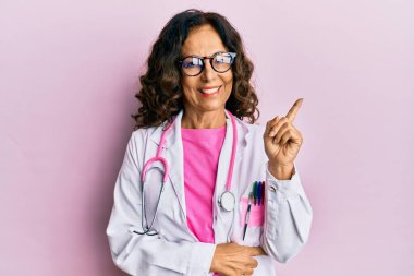 Orta yaşlı İspanyol kadın doktor üniforması giyiyor ve yüzünde kocaman bir gülümseme olan gözlük takıyor. Kameraya el ve parmak şeklinde bakıyor.. 