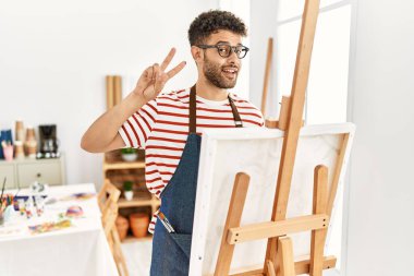 Sanat stüdyosundaki Arap genç adam barış sembolü yapıyor. Yüzünde parmaklar, gülümseyerek zaferi gösteriyor. 