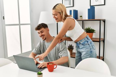 Genç beyaz çift dizüstü bilgisayar kullanıyor ve kahve içiyor. Evdeki masada oturuyorlar..