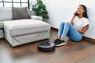 Akıllı telefondan konuşan genç Latin kadın evde akıllı robot süpürgesi kullanıyor.