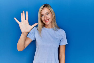 Güzel sarışın kadın mavi arka planda rahat bir tişört giyer ve parmaklarıyla beş numarayı gösterirken kendinden emin ve mutlu bir şekilde gülümserdi.. 